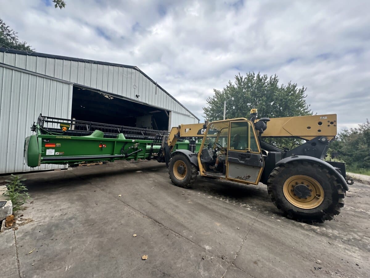 Yellow CAT telehandler carrying a green John Deere header out of a pole barn