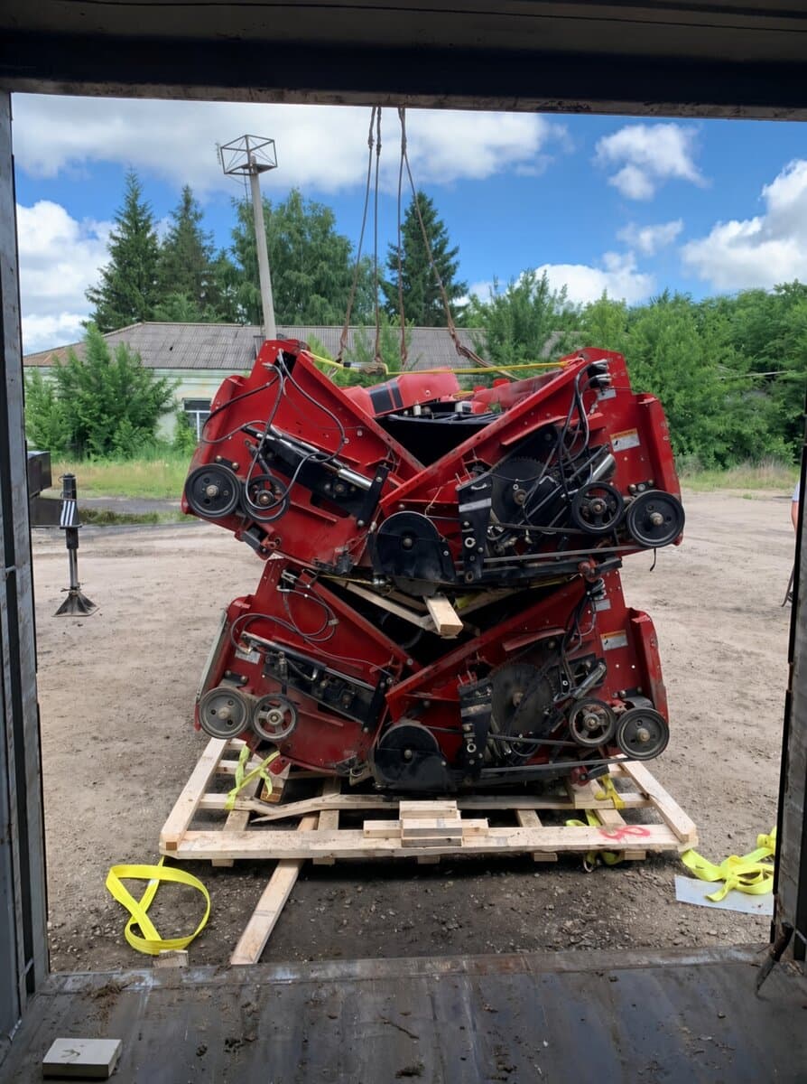 Two red combine header assemblies suspended by crane straps over a wooden pallet at a loading bay