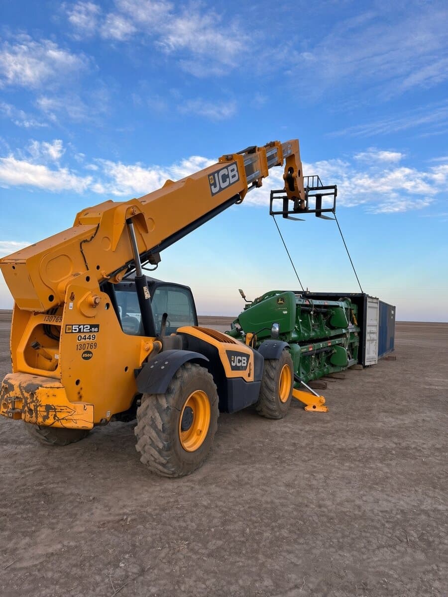 Yellow JCB telehandler lifting a green John Deere header toward an open shipping container